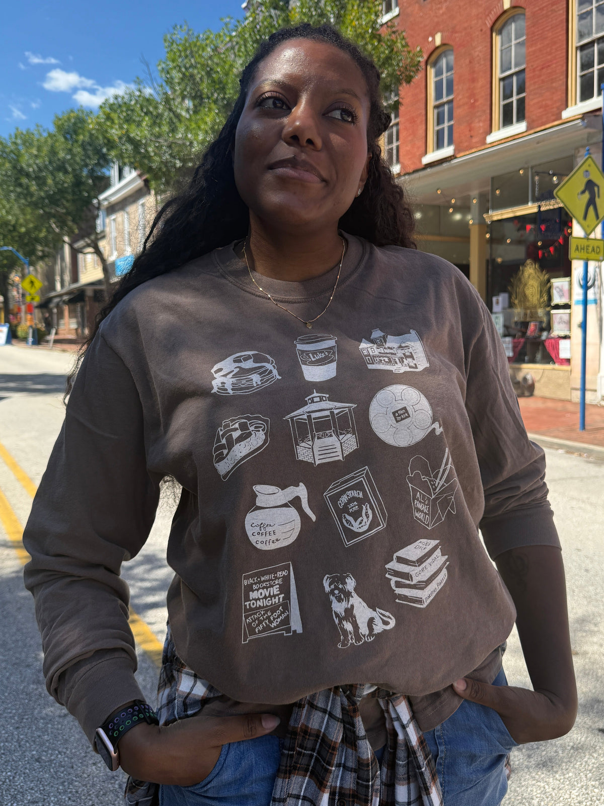 A woman standing on a city street wearing a brown long-sleeve graphic sweatshirt with white illustrations of coffee cups, books, a dog, and other objects, paired with a plaid shirt tied at the waist and blue jeans.